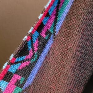 K&ouml;nnte beinhalten: Nahaufnahme eines braunen gewebten Textils mit einem farbenfrohen geometrischen Muster. Das Muster umfasst rosa, blaue, gr&uuml;ne und schwarze Formen. Der Stoff hat ein strukturiertes Aussehen.