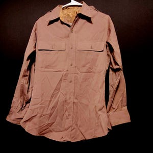 May include: A brown button-down shirt with two chest pockets and long sleeves. The shirt has a collared neckline and is hanging on a white hanger. The shirt is made of a smooth fabric.