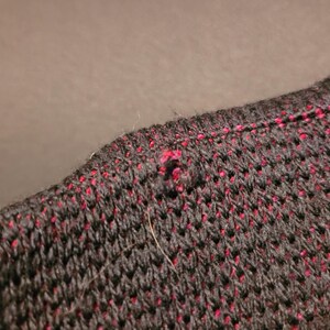K&ouml;nnte beinhalten: Nahaufnahme eines Abschnitts eines Strickkleidungsst&uuml;cks. Der Stoff ist &uuml;berwiegend schwarz mit kleinen, gleichm&auml;&szlig;ig verteilten roten Flecken. Die Textur scheint offen gewebt zu sein, mit einem sichtbaren Loch und ein paar losen Fasern.