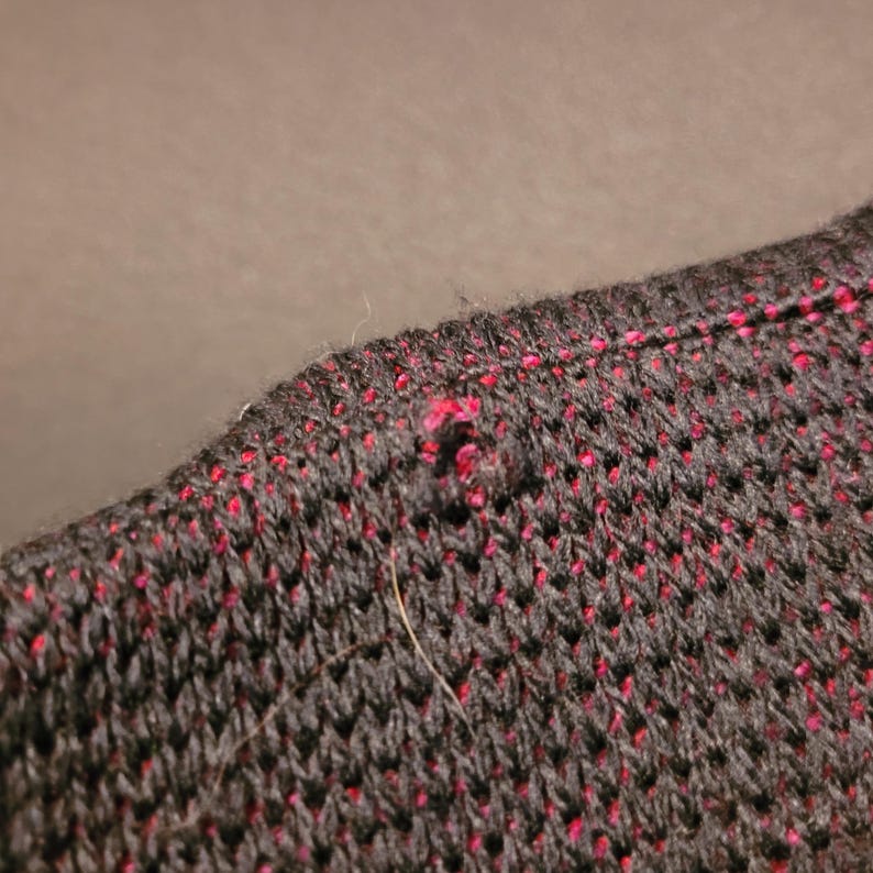 K&ouml;nnte beinhalten: Nahaufnahme eines Abschnitts eines Strickkleidungsst&uuml;cks. Der Stoff ist &uuml;berwiegend schwarz mit kleinen, gleichm&auml;&szlig;ig verteilten roten Flecken. Die Textur scheint offen gewebt zu sein, mit einem sichtbaren Loch und ein paar losen Fasern.