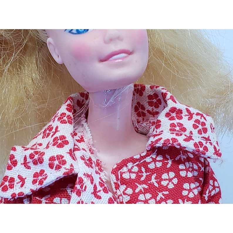 May include: Close-up of a vintage doll's face and upper torso. The doll has blonde hair, blue eyes, and rosy cheeks. She wears a red and white floral print collared shirt. The doll's neck is visible, showing the plastic construction.