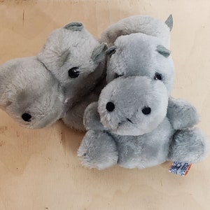 May include: Two gray plush hippopotamus toys. The hippopotamus on the left is lying down with its head turned to the side. The hippopotamus on the right is sitting up with its head facing forward. Both hippopotamuses have black eyes and a small black nose.