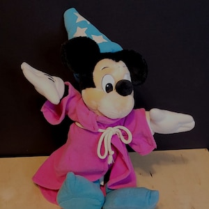 Op de afbeelding: Een pluche Mickey Mouse verkleed als tovenaar. Hij draagt een roze gewaden met een witte touwgordel en een blauwe punthoed met witte sterren. Hij heeft een witte handschoen aan zijn rechterhand en een witte handschoen aan zijn linkerhand.