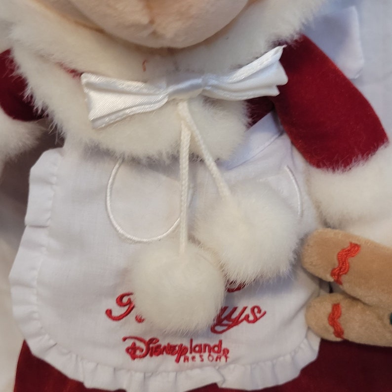 K&ouml;nnte beinhalten: Wei&szlig;es Pl&uuml;schtier mit einem rot-wei&szlig;en Weihnachtsmann-Outfit. Das Spielzeug hat eine wei&szlig;e Schleife, wei&szlig;e Bommeln und eine wei&szlig;e Sch&uuml;rze mit dem gestickten Text "9. Days Disneyland Resort".