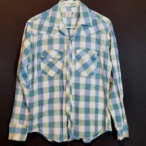Op de afbeelding: Een langarm, geknoopt overhemd met blauw-groen ruitpatroon en twee borstzakken. Het overhemd heeft een puntige kraag en een label met de tekst "Ranger" op de voorkant.