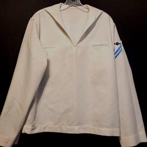 以下が含まれることがあります： 白い長袖シャツ。セーラーカラーと右袖に青と白のパッチが付いている。