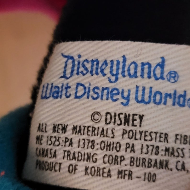 Op de afbeelding: Wit etiket met blauwe tekst die "Disneyland&reg; Walt Disney World &copy;DISNEY ALL NEW MATERIALS POLYESTER FIBE ME 1525:PA 1378:OHIO PA 1378:MASS CANASA TRADING CORP. BURBANK, CA, PRODUCT OF KOREA MFR-100" luidt.