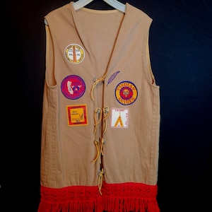May include: Tan vest with multiple patches, including "Indian Guides" and "Calumet Nation." The vest has a red fringe at the bottom and leather ties down the front. Patches feature various colors and designs.