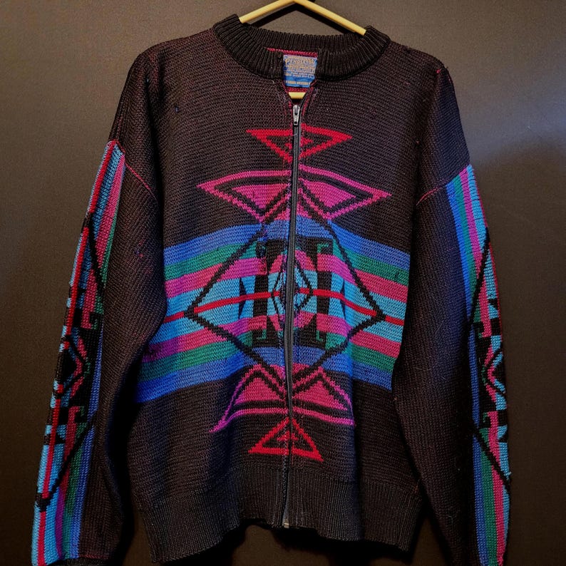 K&ouml;nnte beinhalten: Dunkelbraune Strickjacke mit Rei&szlig;verschluss und geometrischem Muster in Rosa, Blau, Gr&uuml;n und Rot. Die Strickjacke hat einen gerippten Kragen und B&uuml;ndchen, und das Muster wiederholt sich auf den &Auml;rmeln. Der Rei&szlig;verschluss ist silberfarben.