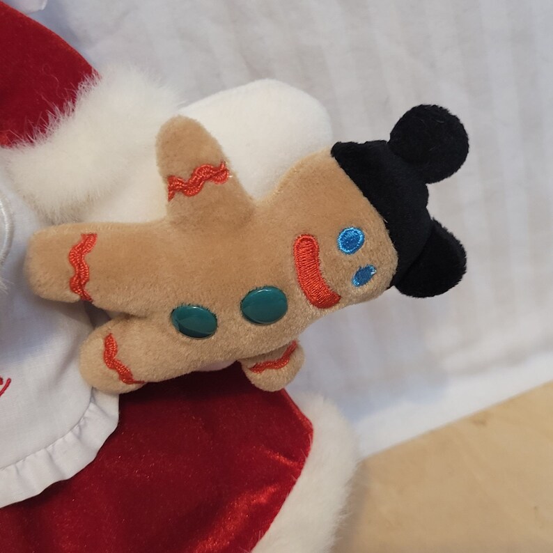K&ouml;nnte beinhalten: Eine Pl&uuml;sch-Weihnachtsmann-Puppe, die ein Lebkuchenmann-Pl&uuml;schtier mit einem schwarzen Mickey-Mouse-Hut h&auml;lt. Der Lebkuchenmann hat gr&uuml;ne Kn&ouml;pfe und einen roten l&auml;chelnden Mund.