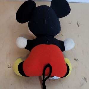 Peut inclure: Un jouet en peluche Mickey Mouse noir et rouge avec une queue noire. Le jouet est assis sur une surface en bois.