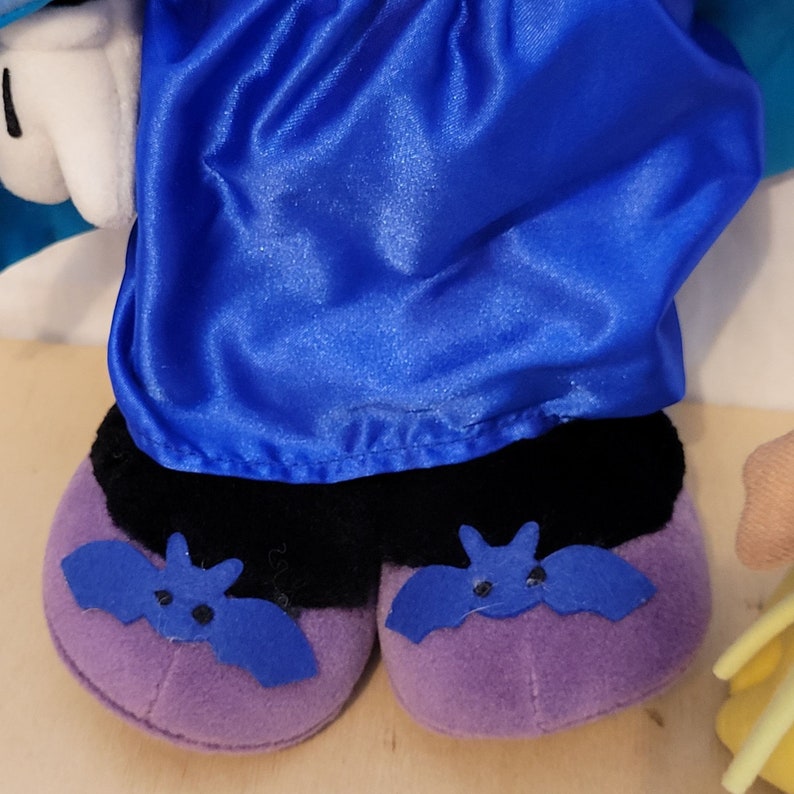 K&ouml;nnte beinhalten: Ein lila Pl&uuml;schtier mit schwarzen F&uuml;&szlig;en und blauen Fledermausfl&uuml;geln. Das Spielzeug tr&auml;gt einen blauen Umhang.