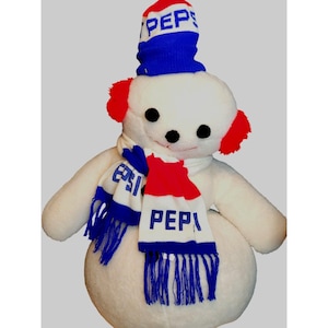 Puede incluir: Un muñeco de nieve de peluche blanco que lleva un gorro a rayas azul y rojo con la palabra "PEPSI" en él. El muñeco de nieve también lleva una bufanda a rayas roja, blanca y azul con la palabra "PEPSI" en ella.