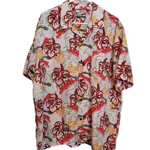 May include: White button-down shirt with a vibrant pattern of red, yellow, and gray floral and abstract designs. The shirt has short sleeves and a collared neckline. The shirt is hanging on a white hanger.