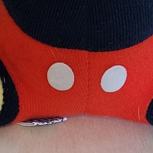 Peut inclure: Peluche rouge avec des oreilles noires et deux cercles blancs sur le devant. Le jouet est fait de tissu doux et a une couture cousue au milieu.