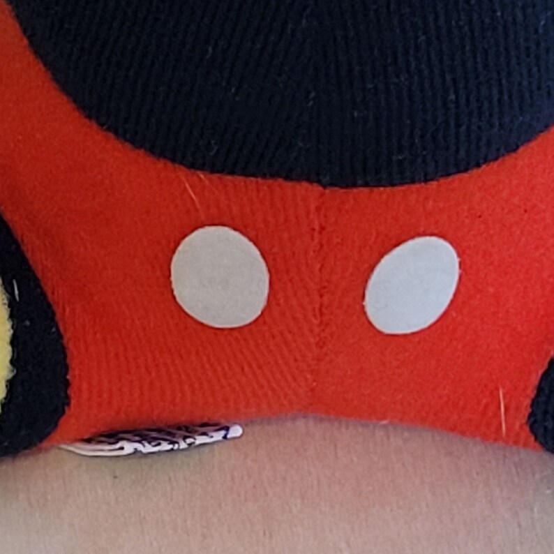 Peut inclure: Peluche rouge avec des oreilles noires et deux cercles blancs sur le devant. Le jouet est fait de tissu doux et a une couture cousue au milieu.