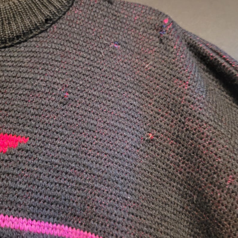 K&ouml;nnte beinhalten: Nahaufnahme eines dunkelgrauen Pullovers mit geripptem Kragen und strukturiertem Strickmuster. Der Pullover hat ein Design aus rosa und magenta geometrischen Formen. Kleine rosa und magentafarbene Flecken sind &uuml;ber den Stoff verstreut.