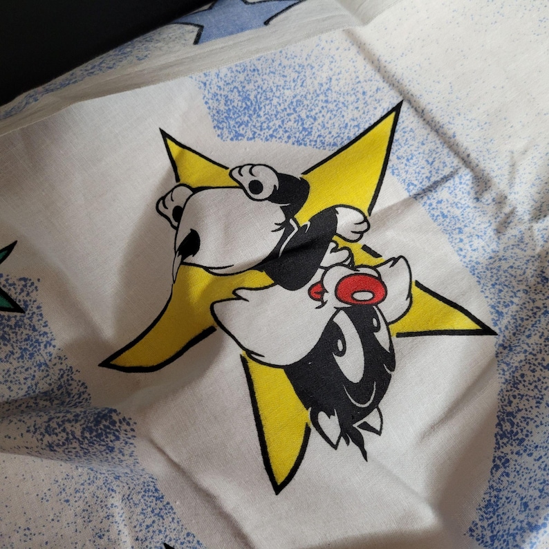 May include: A white fabric with a yellow star and a black and white cartoon character. The character is a bird with large eyes and a red beak. The character is riding on the star.