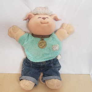 May include: A plush dog doll with a light brown face, ears, and paws. It wears a teal shirt, denim shorts, and a brown collar with a gold pendant. A white cat patch is on the shirt. The doll has light brown hair.