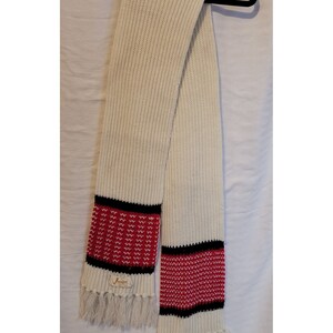 May include: Cream-colored knitted scarf with red and black patterned ends and fringe. The scarf is made of a thick, textured yarn. The ends feature a red zig-zag pattern between black stripes. A small label is sewn on the end.