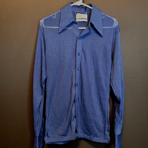 Puede incluir: Una camisa azul de manga larga con botones y cuello. La camisa tiene una tela ligeramente texturizada y un ajuste holgado.