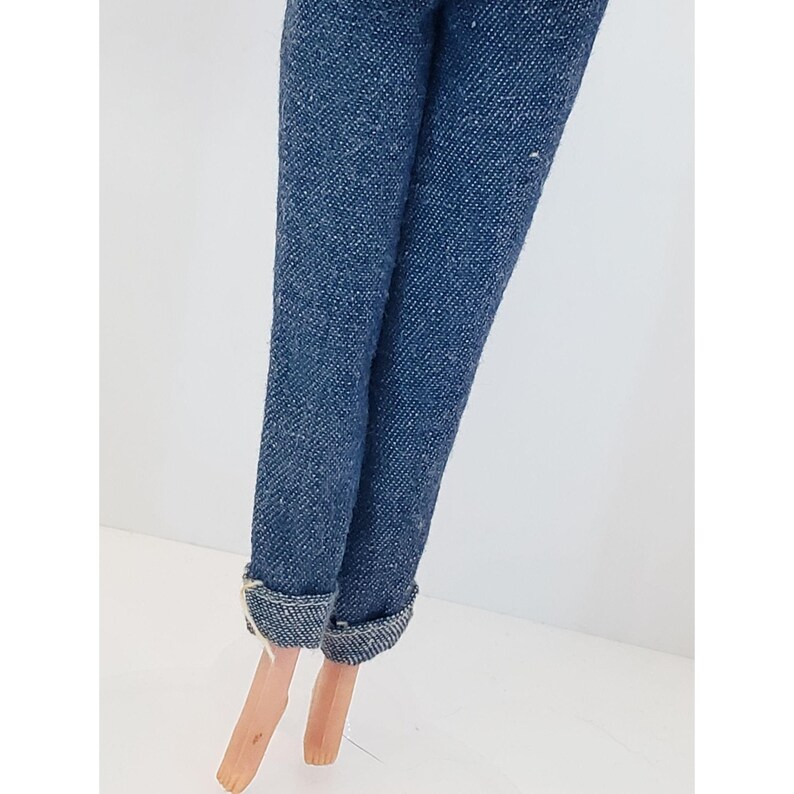 May include: Close-up of doll legs wearing blue denim jeans with cuffed hems. The doll's legs are a light skin tone, and the jeans have a classic, slightly faded appearance. The background is plain white.
