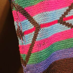 K&ouml;nnte beinhalten: Nahaufnahme eines Stricktextils mit einem lebendigen, farbenfrohen Muster. Das Design zeigt horizontale Streifen in Pink-, Blau-, Gr&uuml;n- und Lilat&ouml;nen, die von diagonalen dunkelbraunen Linien geschnitten werden. Der Strick hat ein strukturiertes Aussehen.