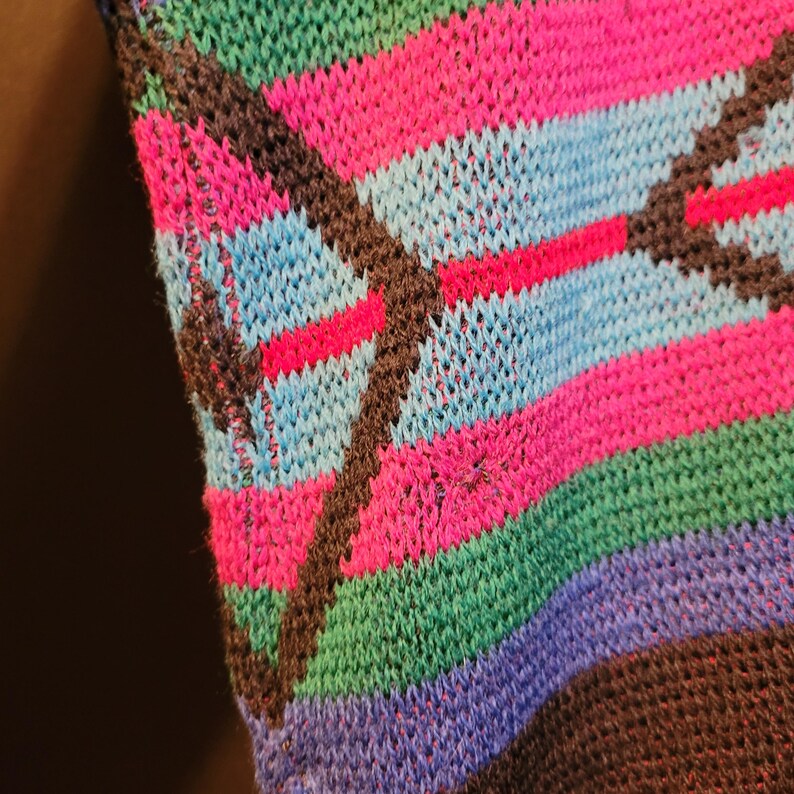 K&ouml;nnte beinhalten: Nahaufnahme eines Stricktextils mit einem lebendigen, farbenfrohen Muster. Das Design zeigt horizontale Streifen in Pink-, Blau-, Gr&uuml;n- und Lilat&ouml;nen, die von diagonalen dunkelbraunen Linien geschnitten werden. Der Strick hat ein strukturiertes Aussehen.