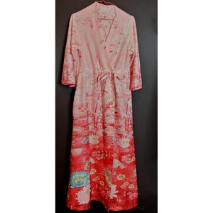 Puede incluir: Una larga bata rosa y roja con estampado floral de nenúfares y juncos. La bata tiene un lazo en la cintura y mangas 3/4.