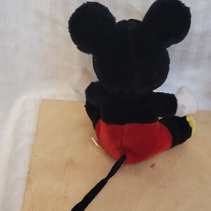 Puede incluir: Un juguete de peluche Mickey Mouse negro y rojo sentado en una superficie de madera. El juguete tiene una cola larga negra y guantes blancos.