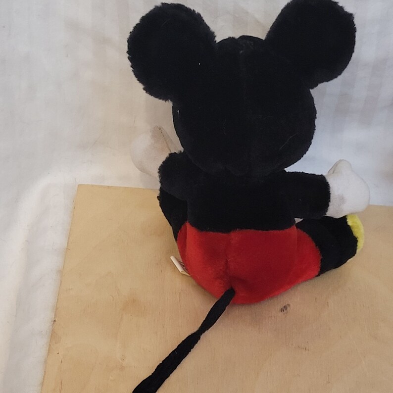 Puede incluir: Un juguete de peluche Mickey Mouse negro y rojo sentado en una superficie de madera. El juguete tiene una cola larga negra y guantes blancos.