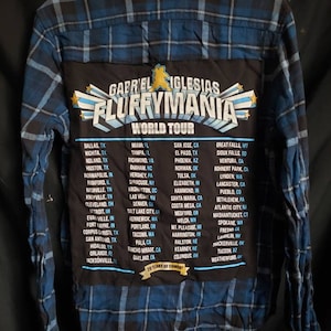 Puede incluir: Una camisa de franela a cuadros azules y negros con un parche negro en la espalda. El parche presenta un oso de dibujos animados y el texto "Gabriel Iglesias Fluffymania World Tour" con una lista de ciudades y estados.