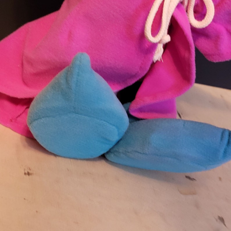 Op de afbeelding: Een pluche speelgoed met een roze capuchon en blauwe benen. Het speelgoed is gemaakt van zacht, pluche stof.