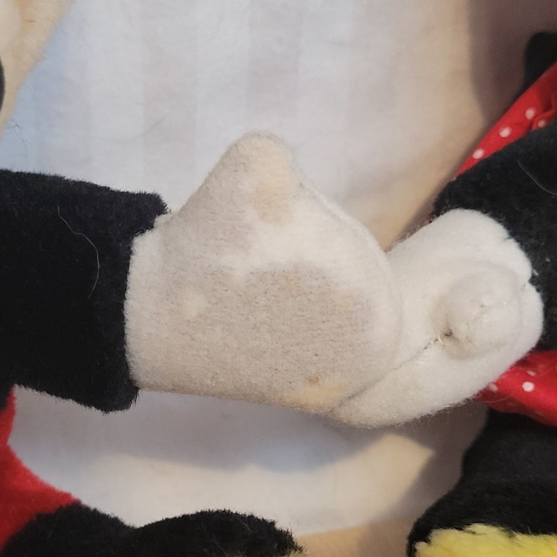 Puede incluir: Primer plano de la mano de un juguete de peluche de Mickey Mouse, que muestra la tela blanca y las costuras. La mano est&aacute; ligeramente sucia.