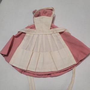 Puede incluir: Un vestido de muñeca rosa y blanco vintage con un delantal plisado y un gorro a juego. El vestido presenta un escote redondo y un delantal plisado. El gorro tiene un borde con volantes y lazos.