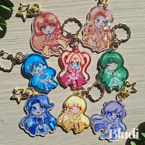 Könnte beinhalten: Eine Sammlung von acht Acryl-Schlüsselanhängern mit Chibi-Anime-Figuren in farbenfrohen Outfits, die jeweils ein Mikrofon halten. Die Anhänger haben eine holografische Oberfläche und sind an goldfarbenen Stern- oder runden Metallverschlüssen befestigt.