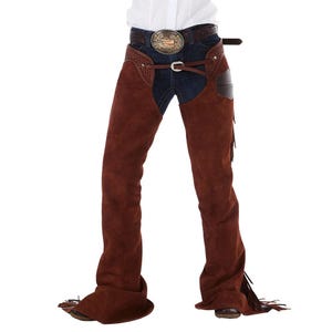 May include: Dark brown suede leather chaps worn over dark blue denim jeans. The chaps feature a textured yoke with decorative stitching, a thin waist belt with a silver buckle, and long fringe along the outer legs. A large oval belt buckle is visible.