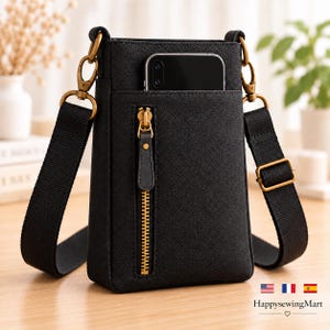 May include: A black textured crossbody phone bag featuring a vertical front zipper pocket with gold-toned hardware, a matching adjustable fabric strap with swivel clips, and a smartphone tucked into the top compartment for storage.