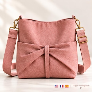 May include: A dusty rose crossbody bag featuring a textured fabric finish and a prominent bow-shaped front pocket detail. The bag includes an adjustable matching shoulder strap attached with antique brass-toned metal swivel clips and hardware.