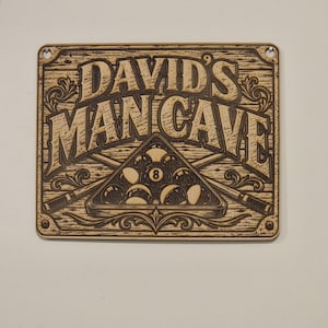 May include: A rectangular wooden sign with rounded corners and two mounting holes, featuring laser-engraved text that reads DAVID'S MAN CAVE above a graphic of a billiard ball rack containing an eight ball and two crossed pool cues.