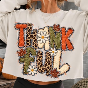 Puede incluir: Una sudadera de manga larga color crema con la palabra THANKFUL en un gráfico texturizado. Las letras incorporan patrones de punto naranja, verde y crema, estampado de leopardo, margaritas blancas y hojas de arce marrones.