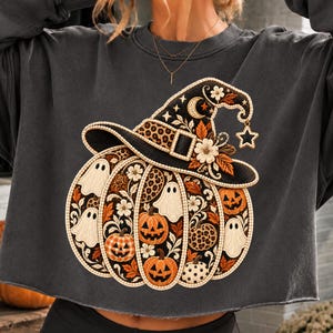 Op de afbeelding: Een antracietgrijs sweatshirt met lange mouwen en een grote geborduurde afbeelding van een pompoen met een heksenhoed. Het ontwerp bevat witte spoken, oranje pompoenen, luipaardprint-secties, bloemmotieven en een ster.