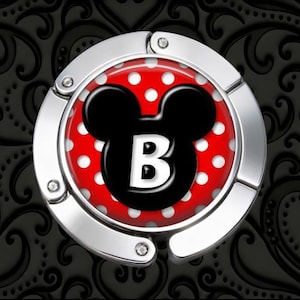 May include: A circular silver-toned metal purse hook featuring a central inlay with a red background and white polka dots. A black silhouette of a mouse head is centered on the inlay, containing a large white capital letter B in the middle.