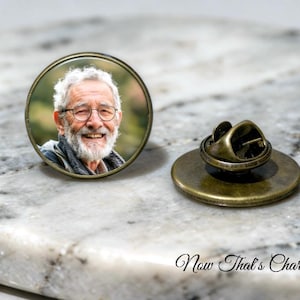 May include: A round metal pin featuring a photographic portrait of an older person with white hair, a beard, and glasses. The pin has a bronze-toned frame and a matching metal clutch backing, displaying a detailed image of a smiling individual.