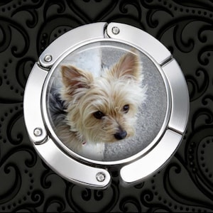 May include: A circular, silver-toned metal bag hanger featuring a central, high-resolution photographic insert of a small, tan and black Yorkshire Terrier dog with pointed ears and dark eyes, set against a black background with a subtle scroll pattern.