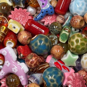 May include: A collection of assorted beads in various shapes, sizes, and materials, including ceramic, glass, and wood. The mix features star, sun, and animal-shaped beads in colours like red, blue, pink, green, and brown with textured finishes.