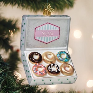 May include: A glass Christmas ornament shaped like an open white box with green polka dots containing six miniature donuts. The inside lid features a pink and teal hexagonal label with the text YUM. The donuts are glazed in brown, gold, and blue.