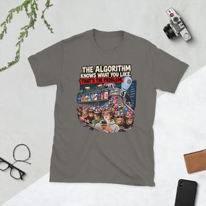 May include: A charcoal gray t-shirt featuring a graphic illustration of a figure labeled The Algorithm pouring various social media icons into the open heads of people looking at smartphones. The text reads: THE ALGORITHM KNOWS WHAT YOU LIKE. THAT'S THE PROBLEM.