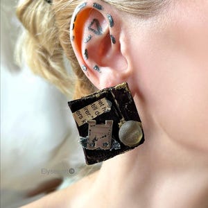 Peut inclure: Une boucle d'oreille carrée avec une base brun foncé, des accents dorés, un morceau de papier avec du texte imprimé, un élément métallique argenté et une perle ronde translucide. La boucle est portée sur une oreille avec des marques argentées.