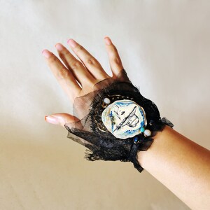 K&ouml;nnte beinhalten: Ein schwarzes Spitzen-Armband mit einem wei&szlig;en Keramik-Anh&auml;nger mit einem blauen und schwarzen Design. Der Anh&auml;nger ist mit goldenen und blauen Perlen verziert.